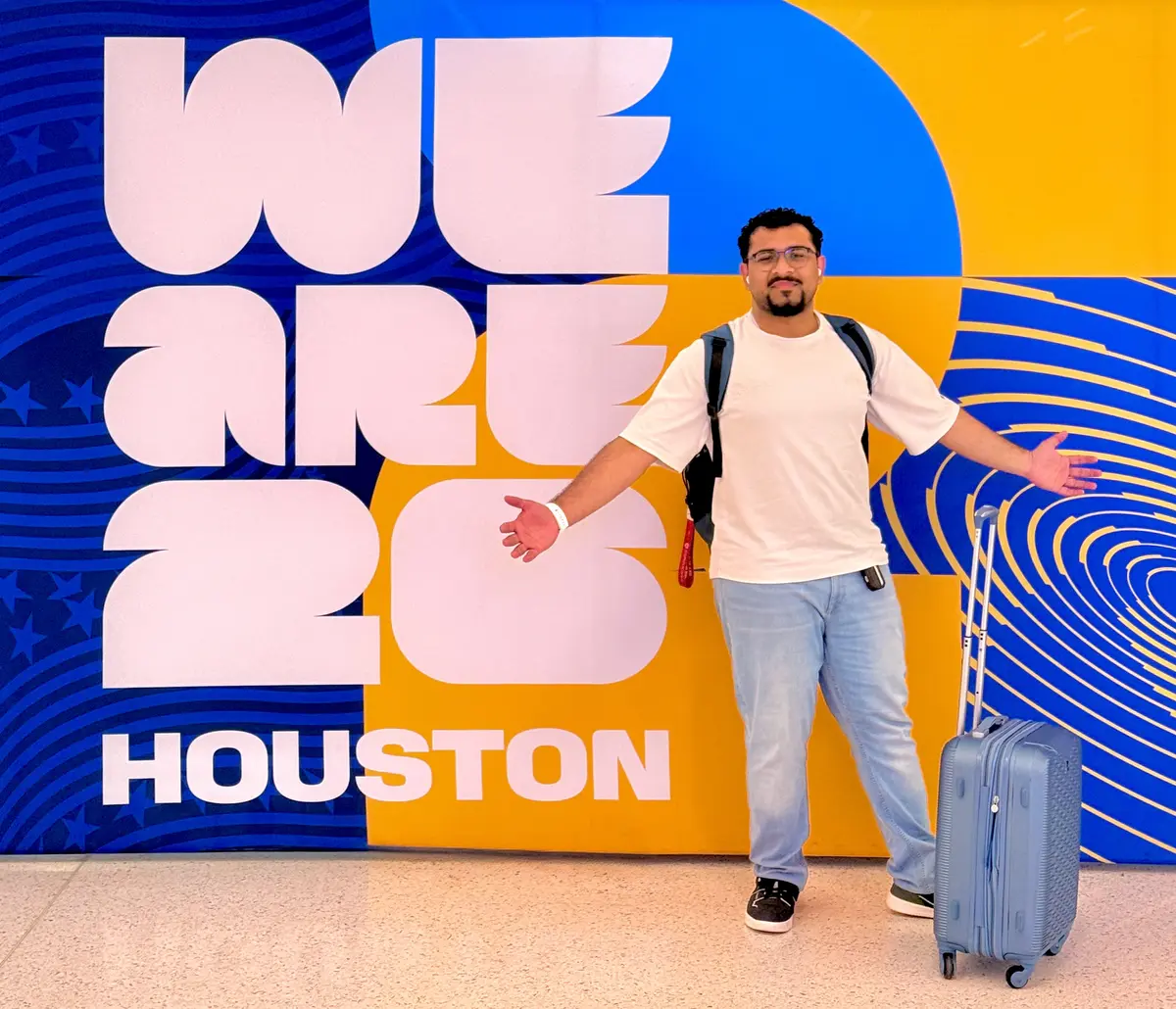 Muhammad Ahmad arriving in Houston beside a colourful airport welcome wall and suitcase.
