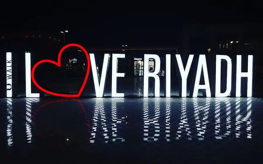 Illuminated I Love Riyadh sign reflected on wet pavement at night.
